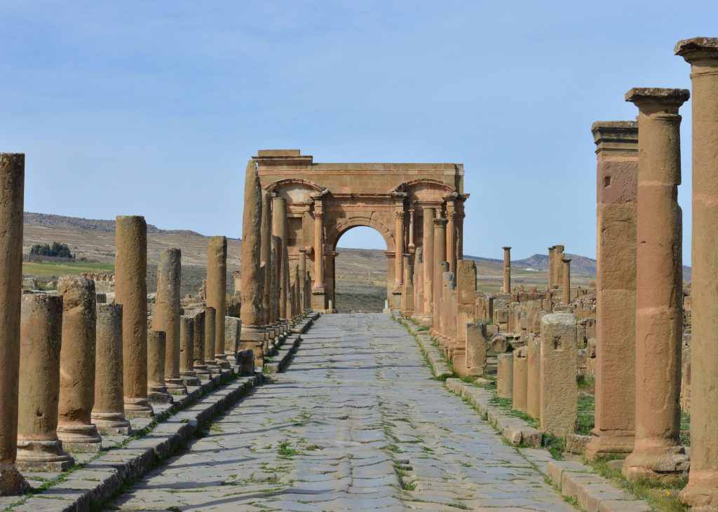 A paved ancient street lined with stone columns leading to a triumphal arch