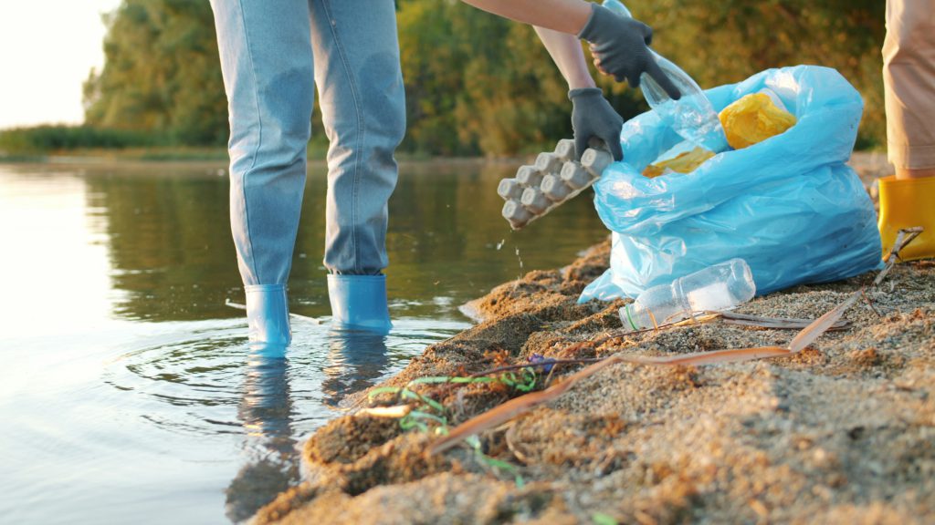 Two people in rubber boots collecting rubbish into a blue bag at the edge of a river